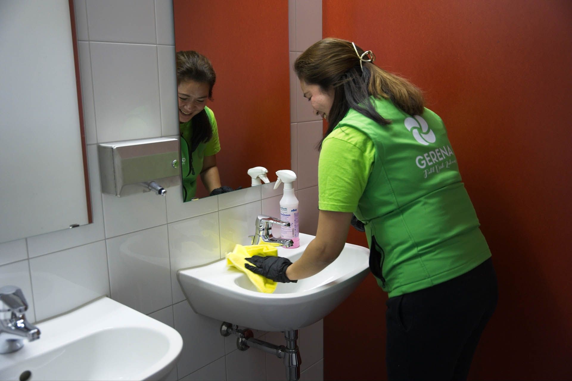 Eine Person in einer grünen Weste reinigt ein Waschbecken im Badezimmer mit einem gelben Tuch.