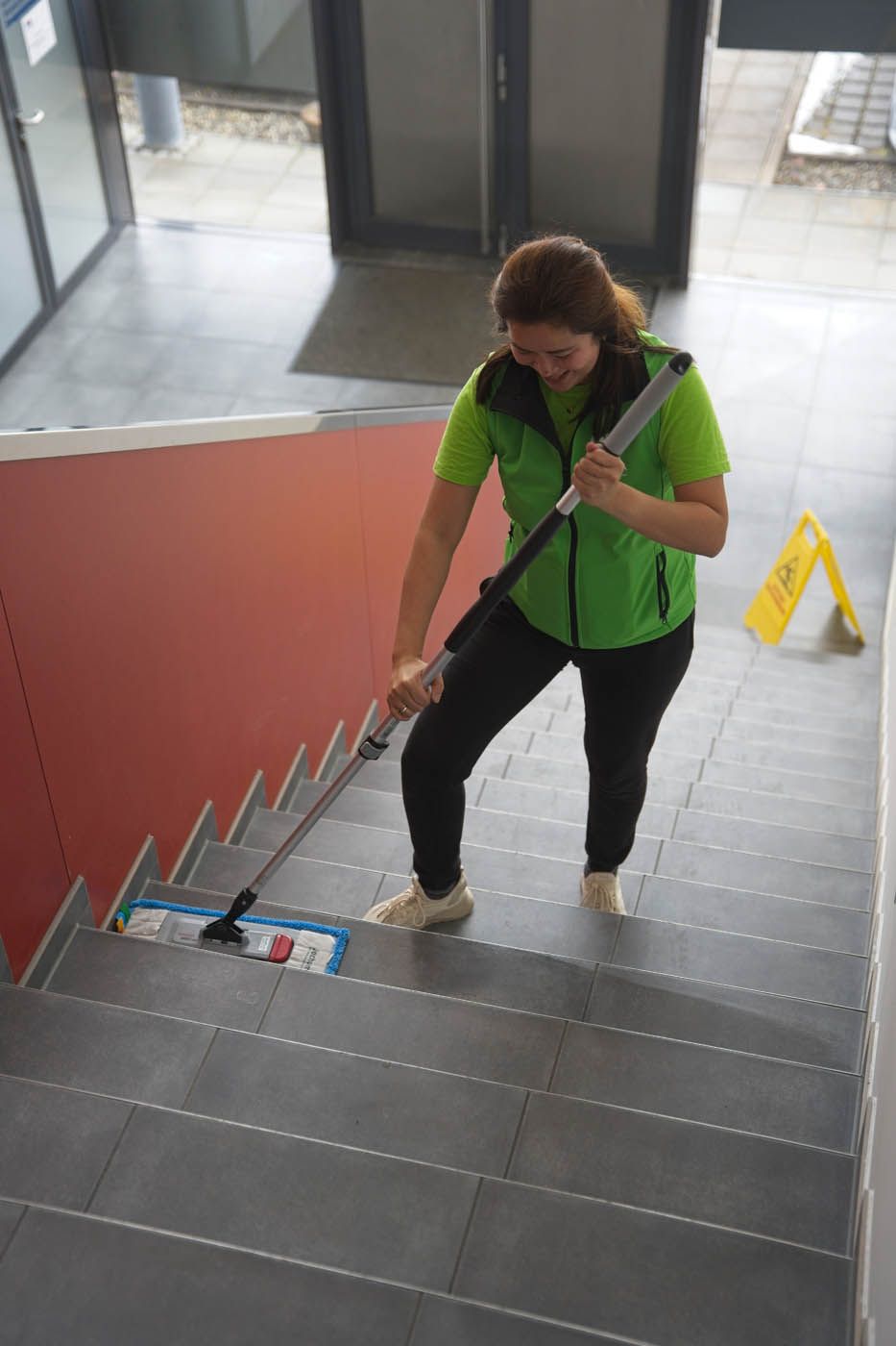 Frau in Grün putzt Treppe mit einem Mopp mit langem Stiel; Warnschild in der Nähe.