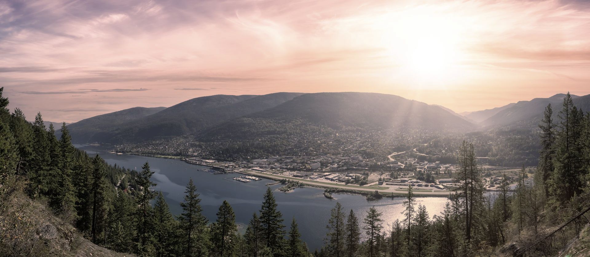 the sun is setting over a lake surrounded by mountains and trees in Nelson, BC