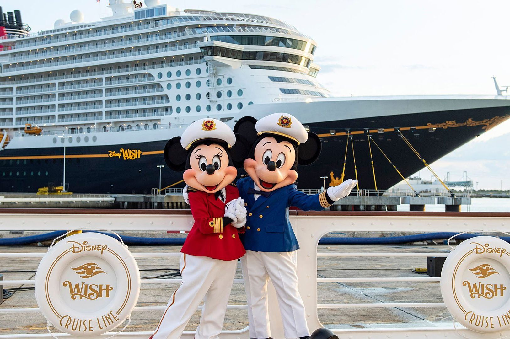 Mickey and Minnie Mouse in captain outfits pose in front of the Disney Wish cruise ship.