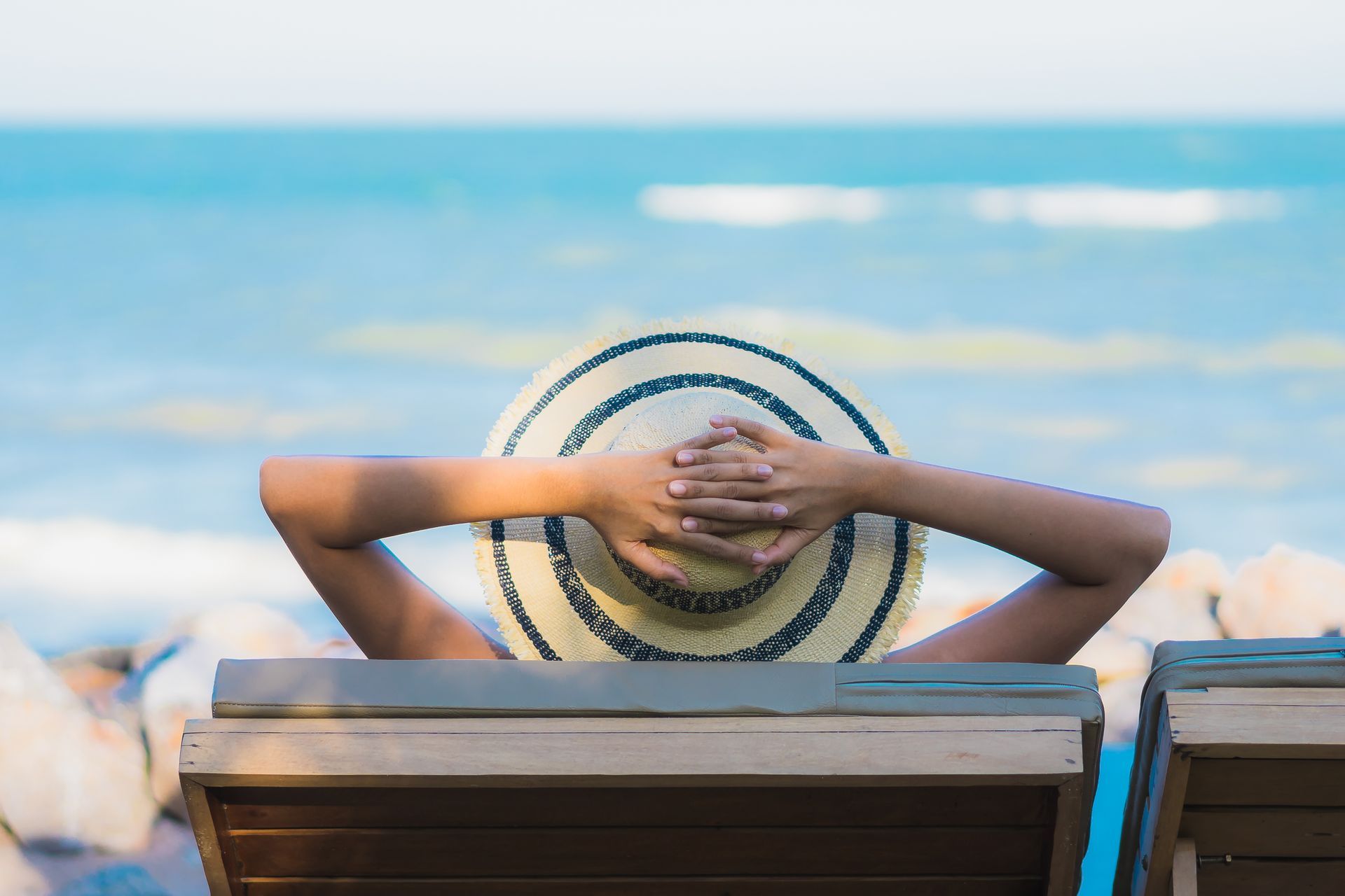Person relaxing on a wooden lounge chair with hands behind head, looking at the ocean.