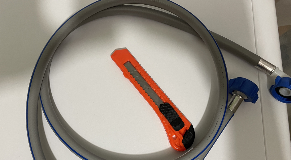 A coiled grey washing machine inlet hose with blue threaded ends, paired with an orange utility knife.