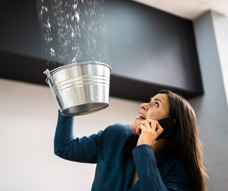 Woman on phone catches water leaking from ceiling into a bucket in a commercial building.