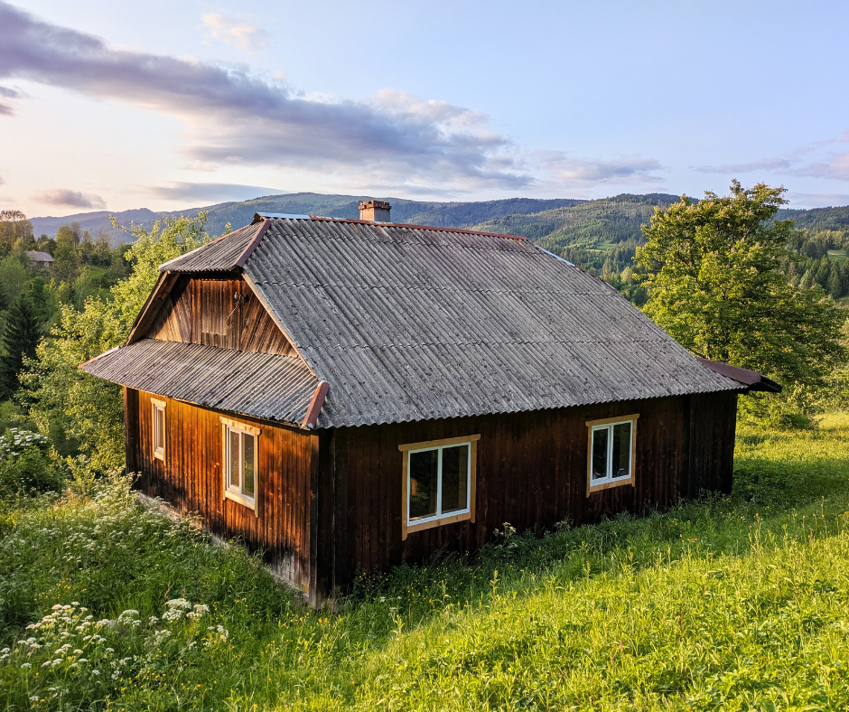 A rustic wooden house with a corrugated roof sits in a grassy, sunlit field before rolling green hills.