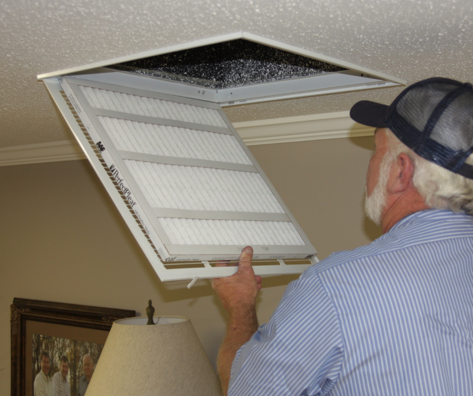 A person with a white beard and hat opening a hinged ceiling HVAC vent to replace or inspect the pleated air filter.