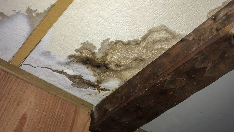 Water-damaged ceiling with brown stains and peeling texture, near a wooden beam and wall.