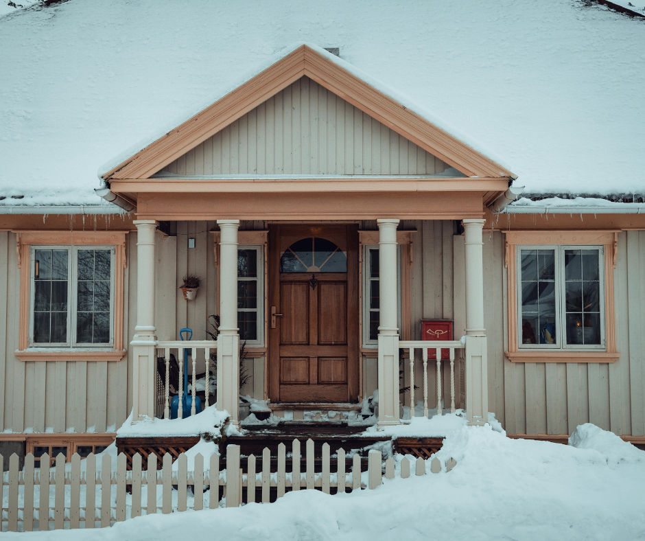 Snow-covered front porch of a beige house with white columns, wooden door, and picket fence