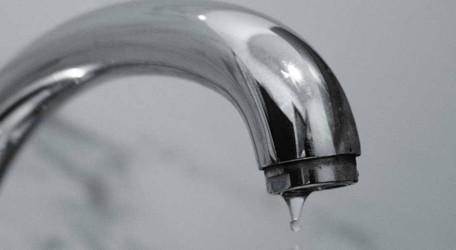 A silver kitchen faucet dripping a single drop of water against a blurred, light background.