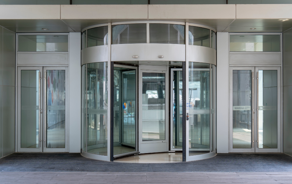 Revolving glass door in a building entrance with two adjacent glass doors. Gray concrete and white framing.
