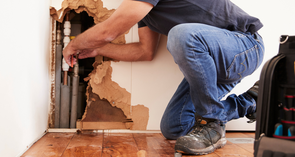 A person kneels on a wooden floor, reaching into a hole in a wall to repair exposed plumbing pipes.