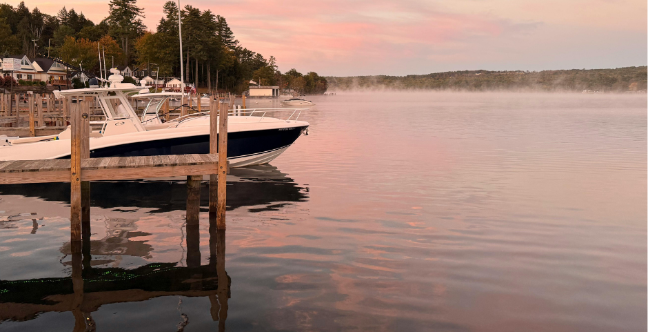 A white boat docked at a wooden pier on a lake during a hazy, pink-hued sunrise.