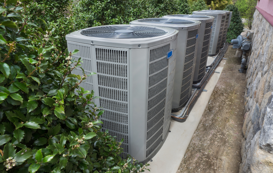 Row of outdoor HVAC units beside a stone wall and hedge on a narrow concrete path