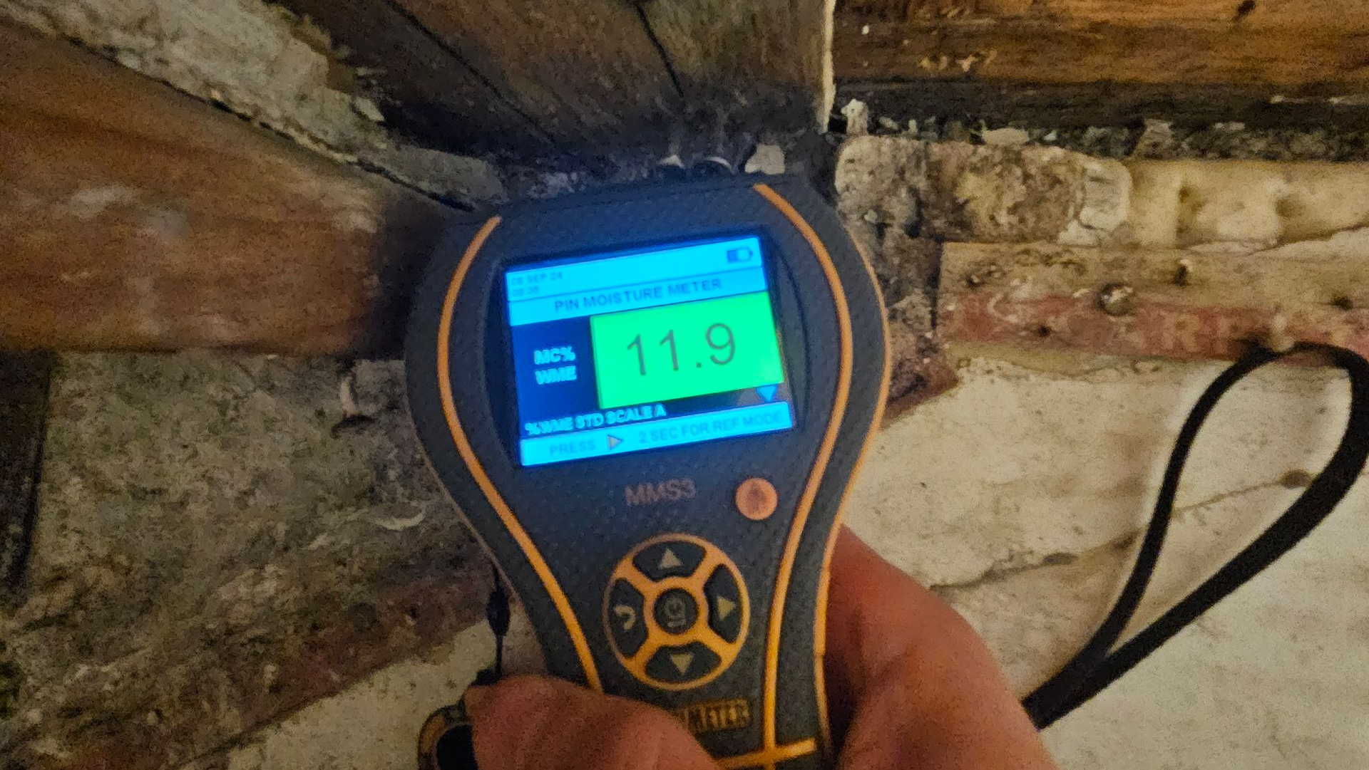 Handheld digital moisture meter reading 11.9 in a stone wall cavity, held near exposed wood beams.