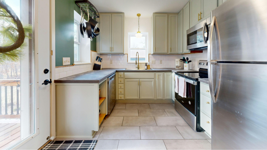 A kitchen with sage green cabinets, light stone floors, stainless steel appliances, and a window above the sink.