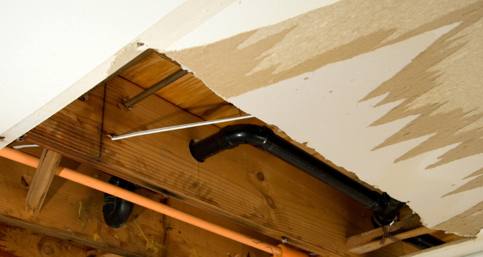 Hole in a ceiling revealing plumbing pipes and wooden supports. Brown and black pipes visible with damaged drywall.