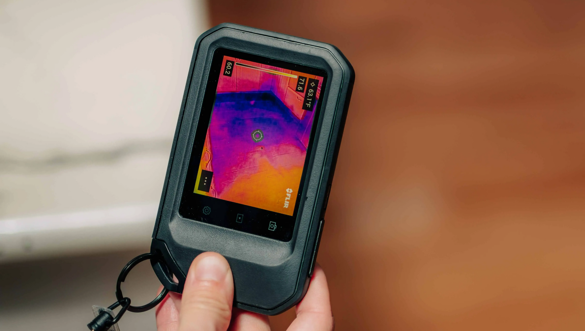 A handheld thermal imaging camera displays a heat map with purple and orange gradients on its screen.