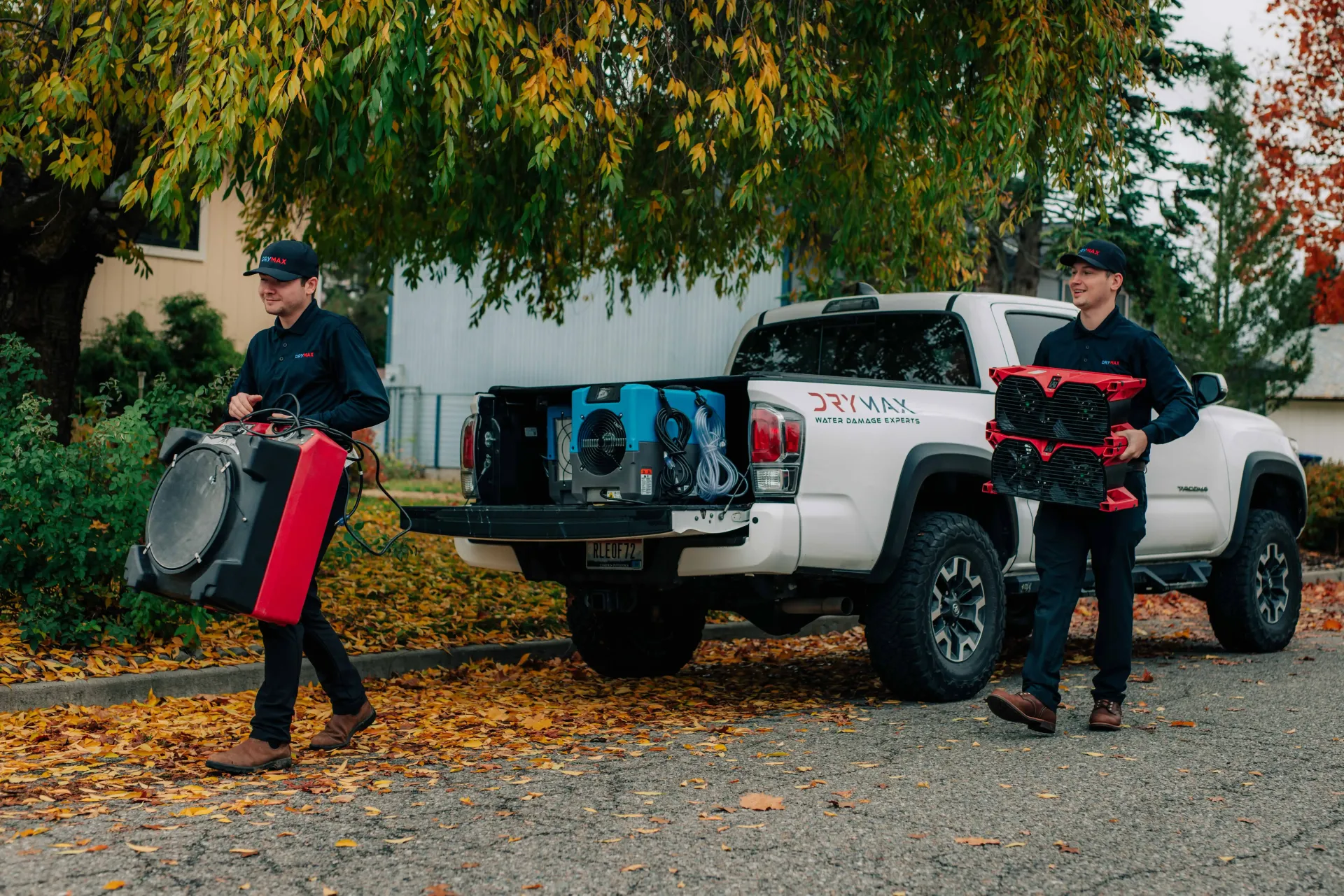 Two workers in black uniforms carry restoration equipment toward a white pickup truck parked on a street with fall leaves.