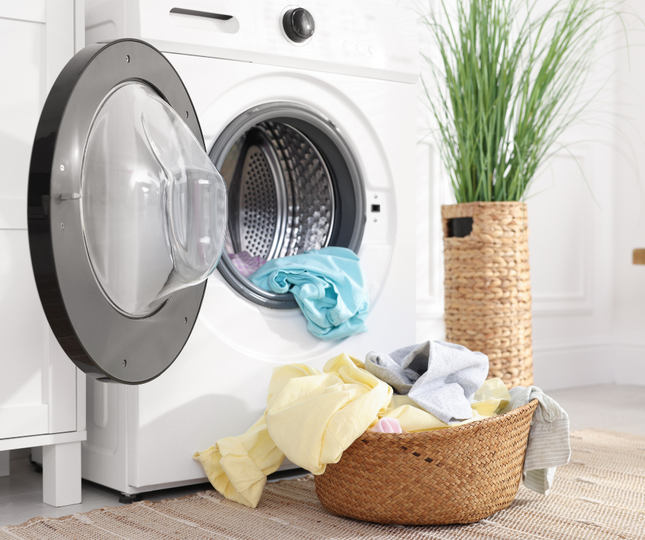 A front-loading white washing machine with an open door, a blue garment inside, and a wicker basket filled with laundry.