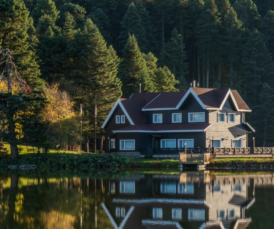 Lakeside cabin with dark wood siding, surrounded by tall green trees, reflected in the water.