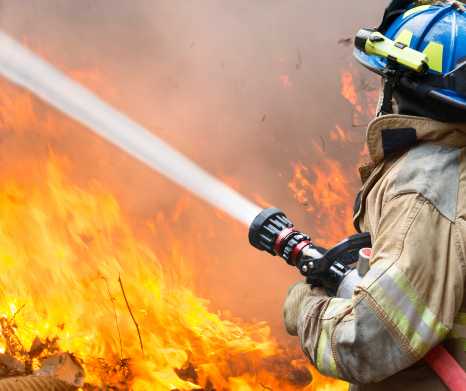 Firefighter aiming a hose at a large, raging fire.