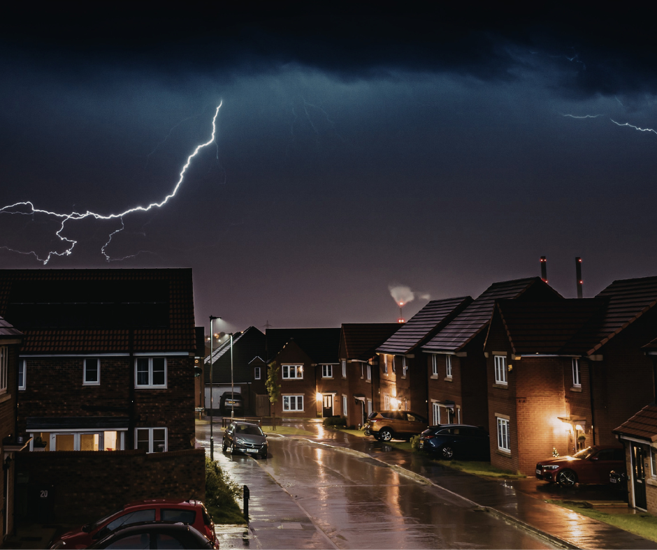 Lightning strikes over a residential street at night, illuminated by streetlights and house lights.