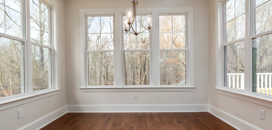 Interior room with multiple white-framed windows overlooking a wooded area. Brown hardwood floor.