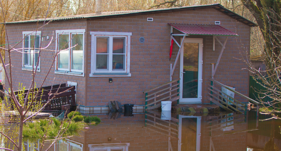 Flooded house with water up to the foundation. Brown siding, white framed windows, and a red awning.