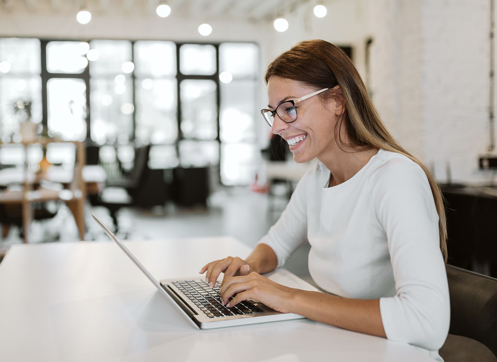 A smiling woman in glasses working on a laptop in a bright office.