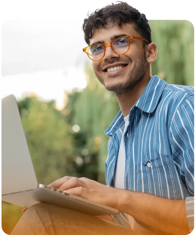 Man wearing orange glasses smiles while using a laptop outdoors.