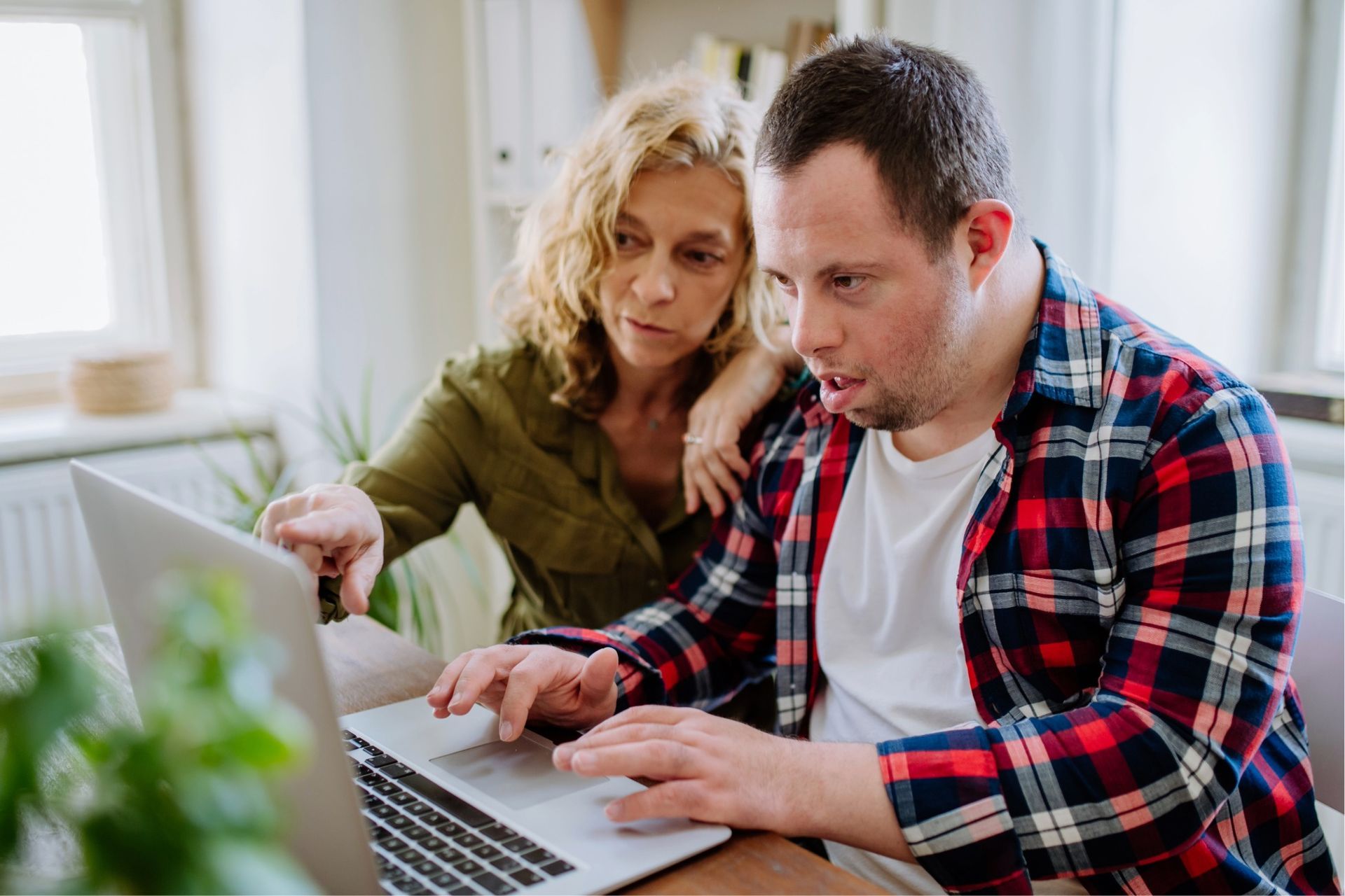 Woman helps a person with Down syndrome use a laptop, pointing at the screen. Indoors.