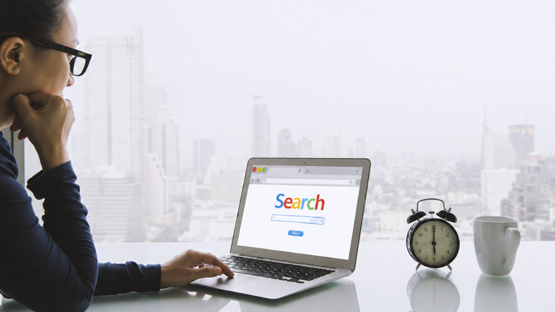 Person working on laptop with search page, beside an alarm clock and coffee mug on a desk