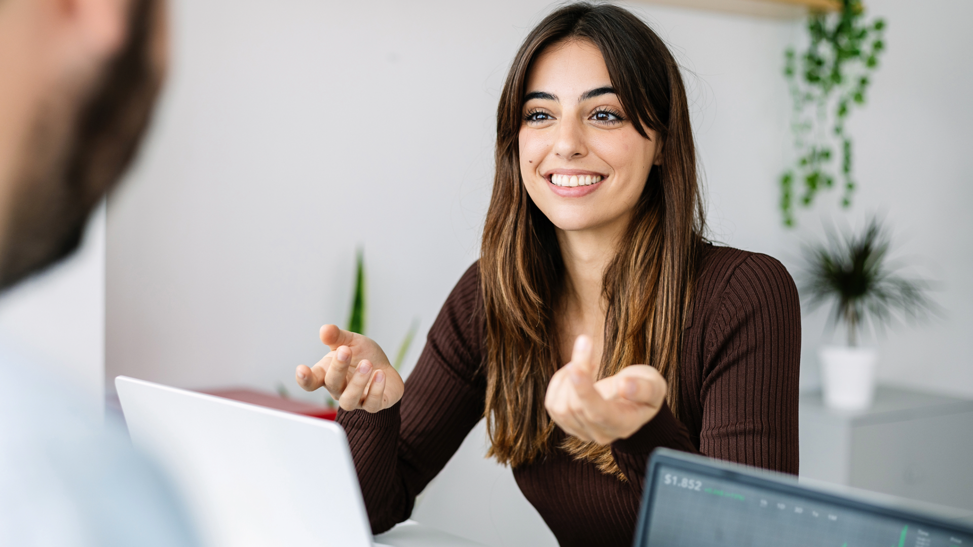 Smiling woman gesturing during a video call in a bright office, with a laptop in the foreground