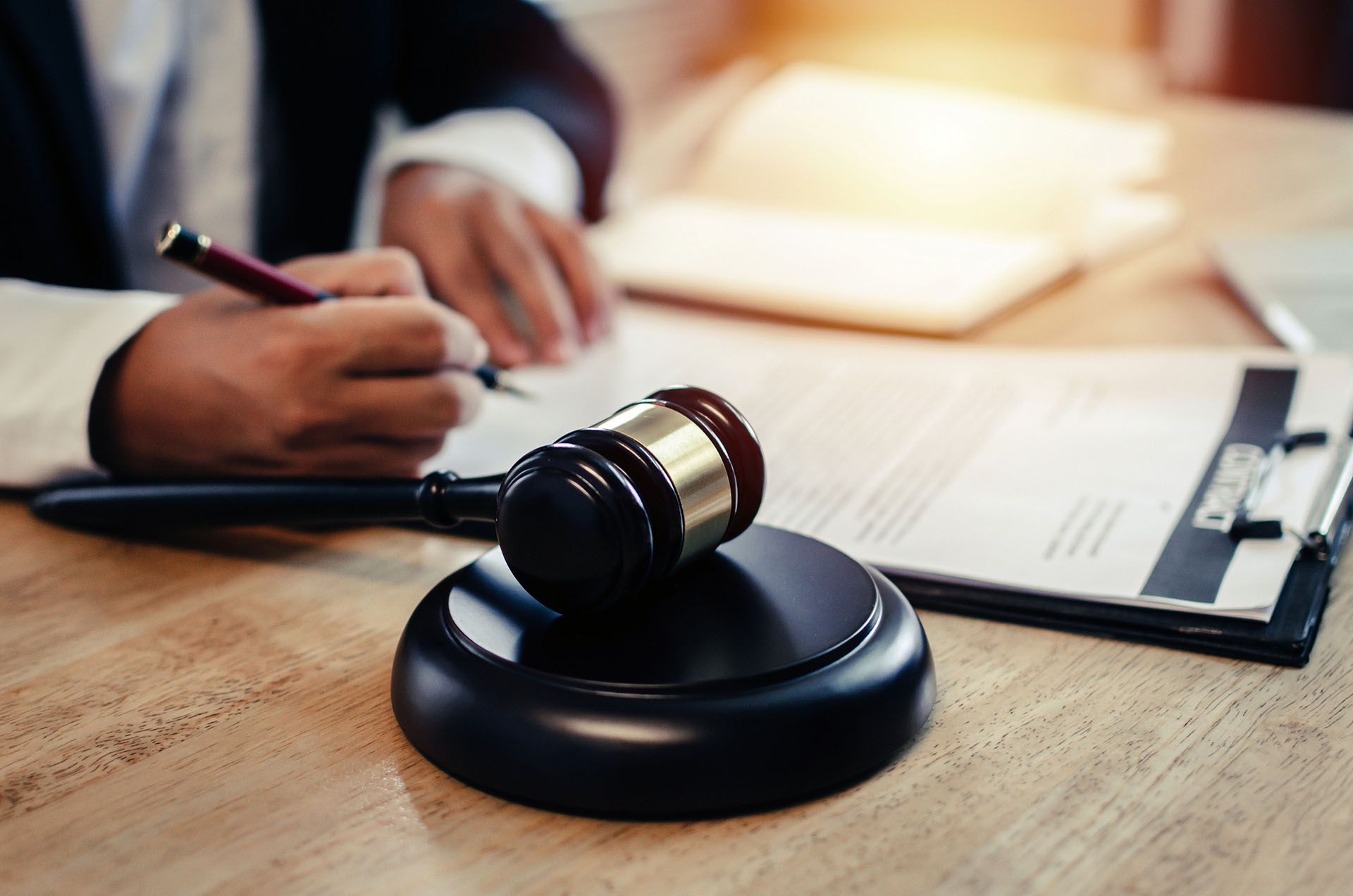 A judge 's gavel is sitting on a wooden table next to a person writing on a clipboard.