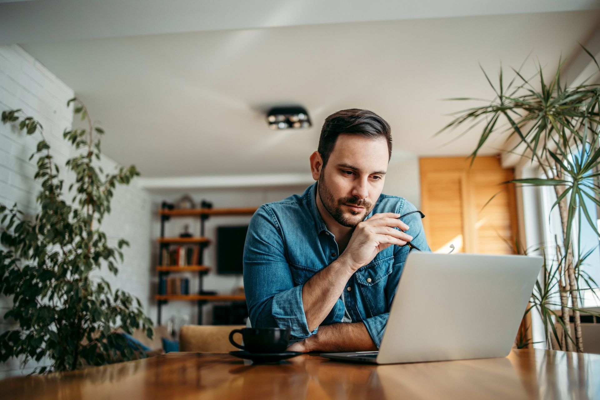 Man in denim shirt, at a table with laptop, indoors, looking at screen, hand to chin, coffee cup nearby.