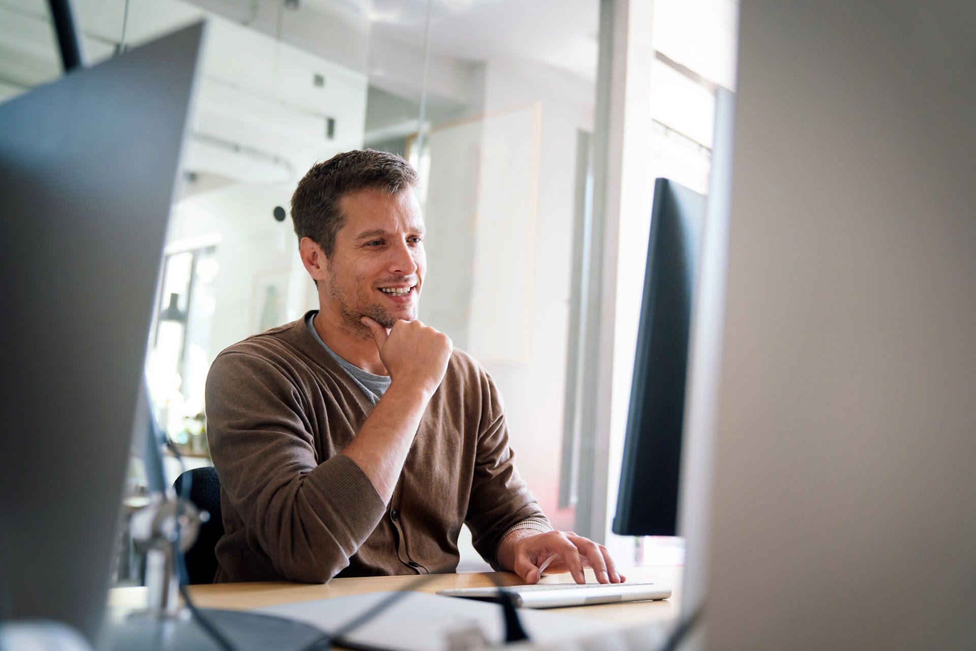 Man in brown sweater smiles while typing at a computer in a bright office.