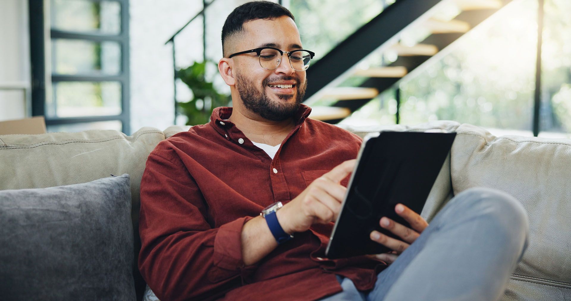Man with glasses smiling, using a tablet while relaxing on a couch.