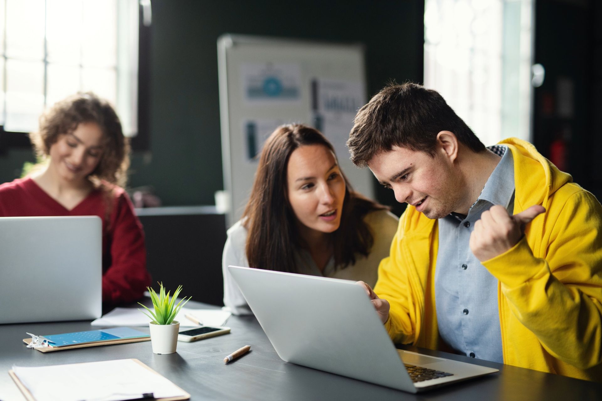 Three people working on laptops at a table. Woman helps man in yellow jacket, he gestures. Another woman works in the background.