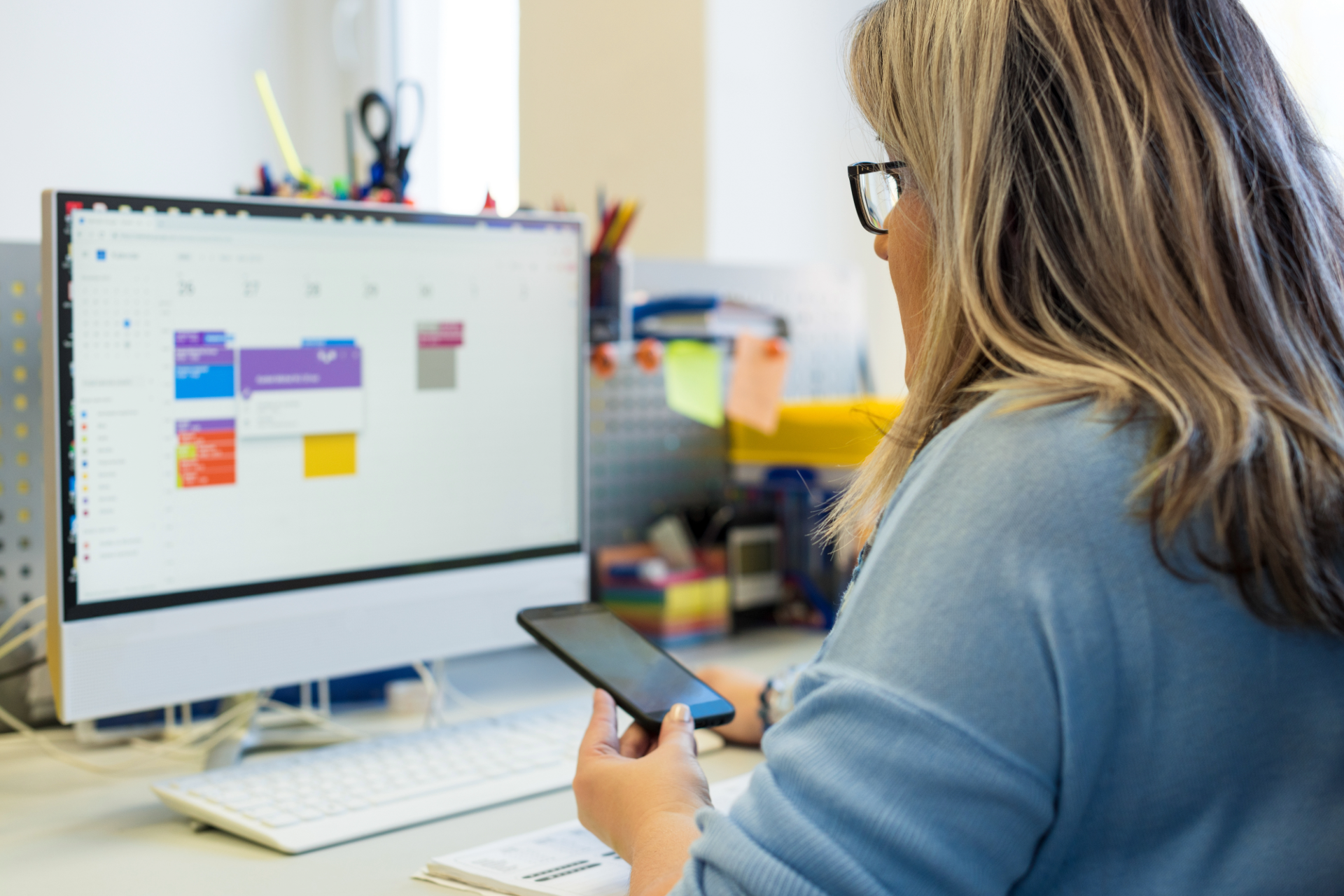 Woman looking at a computer monitor and phone at a desk, reviewing a calendar.