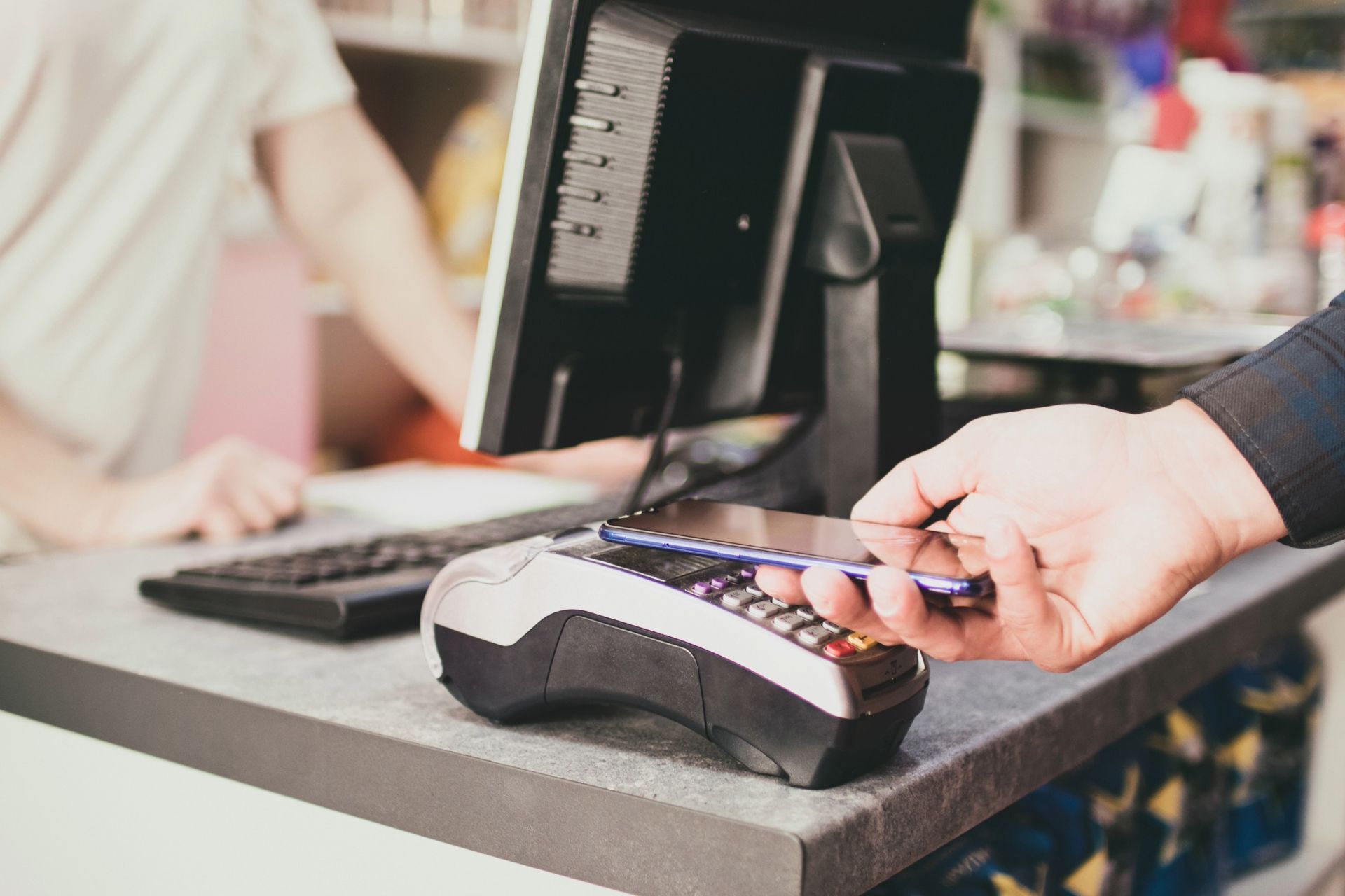 Person using phone to pay at a point-of-sale terminal. A cashier is in the background.
