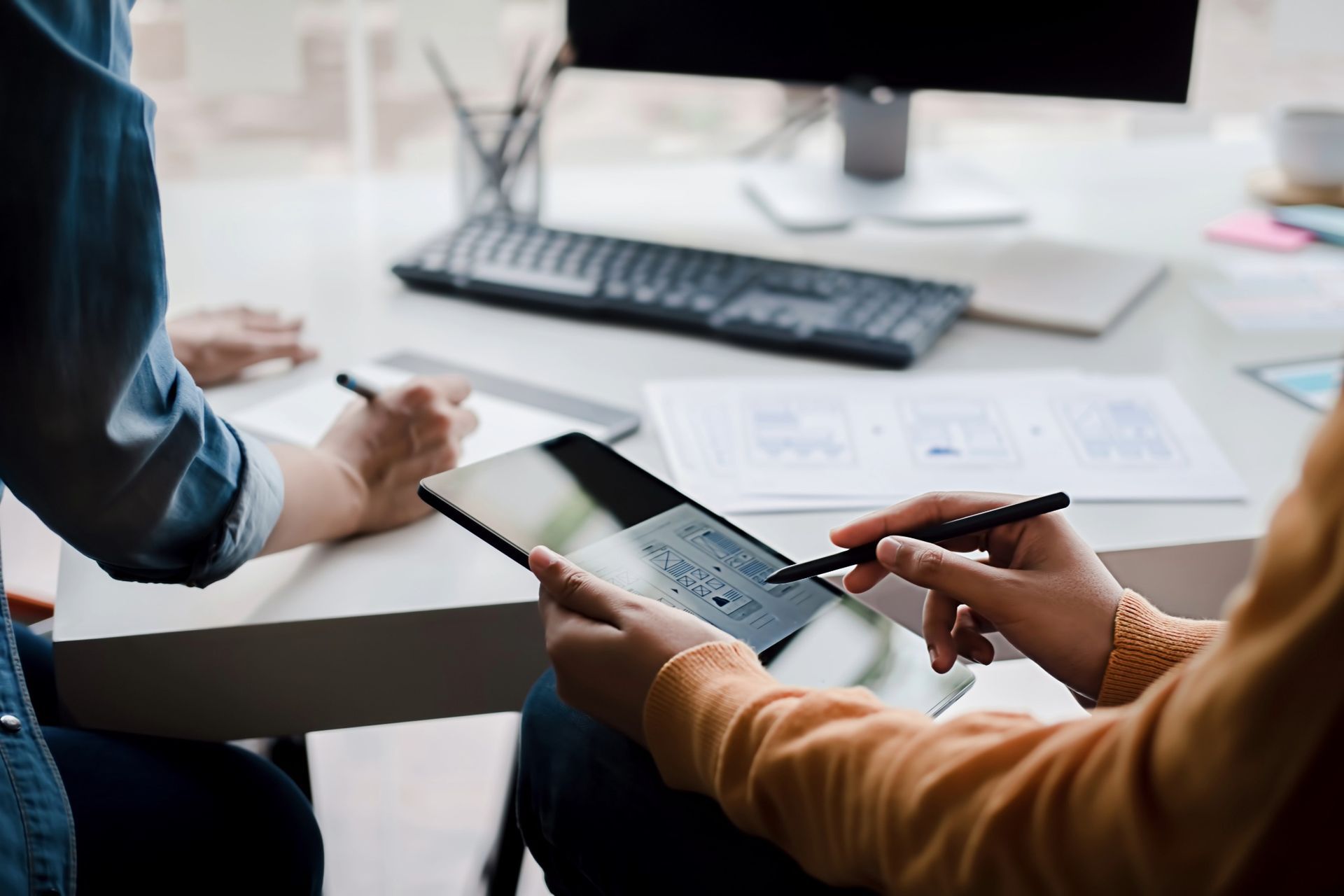 Two people collaborating at a desk, one using a tablet with a stylus, the other writing.