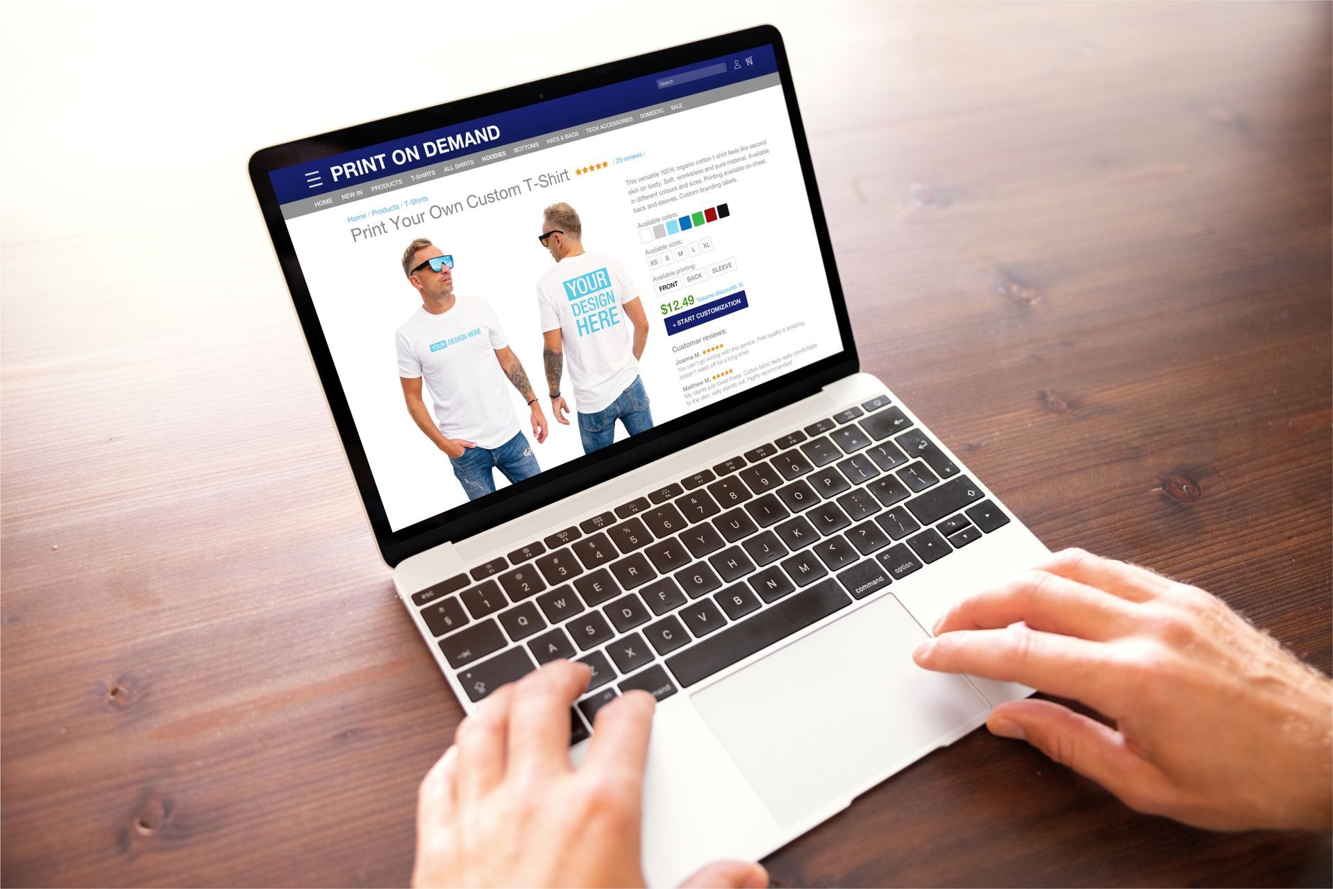 Person using laptop, browsing a t-shirt website with two men modeling shirts on a wood table.