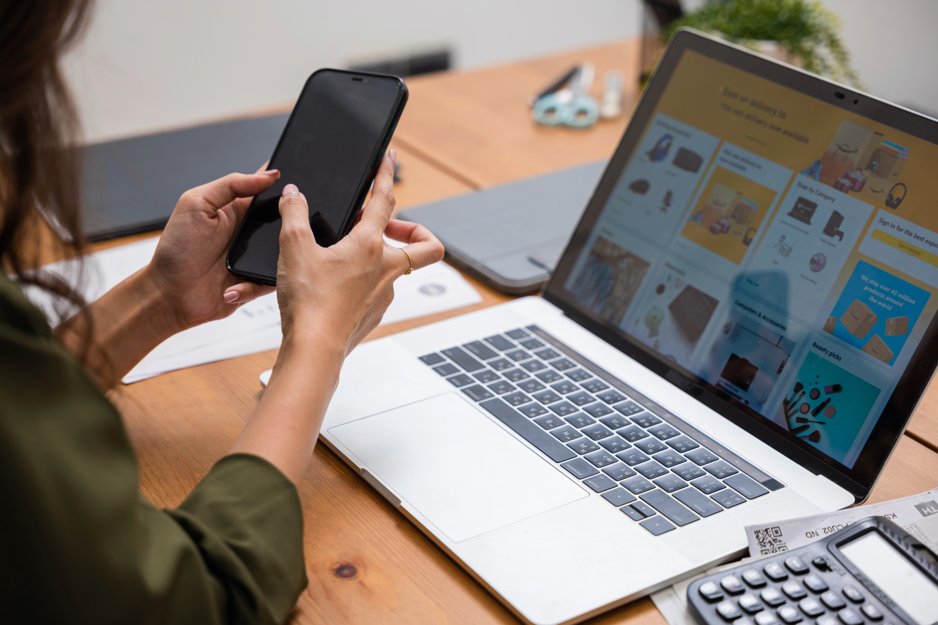 Person using a smartphone and laptop at a desk with a calculator and other items.