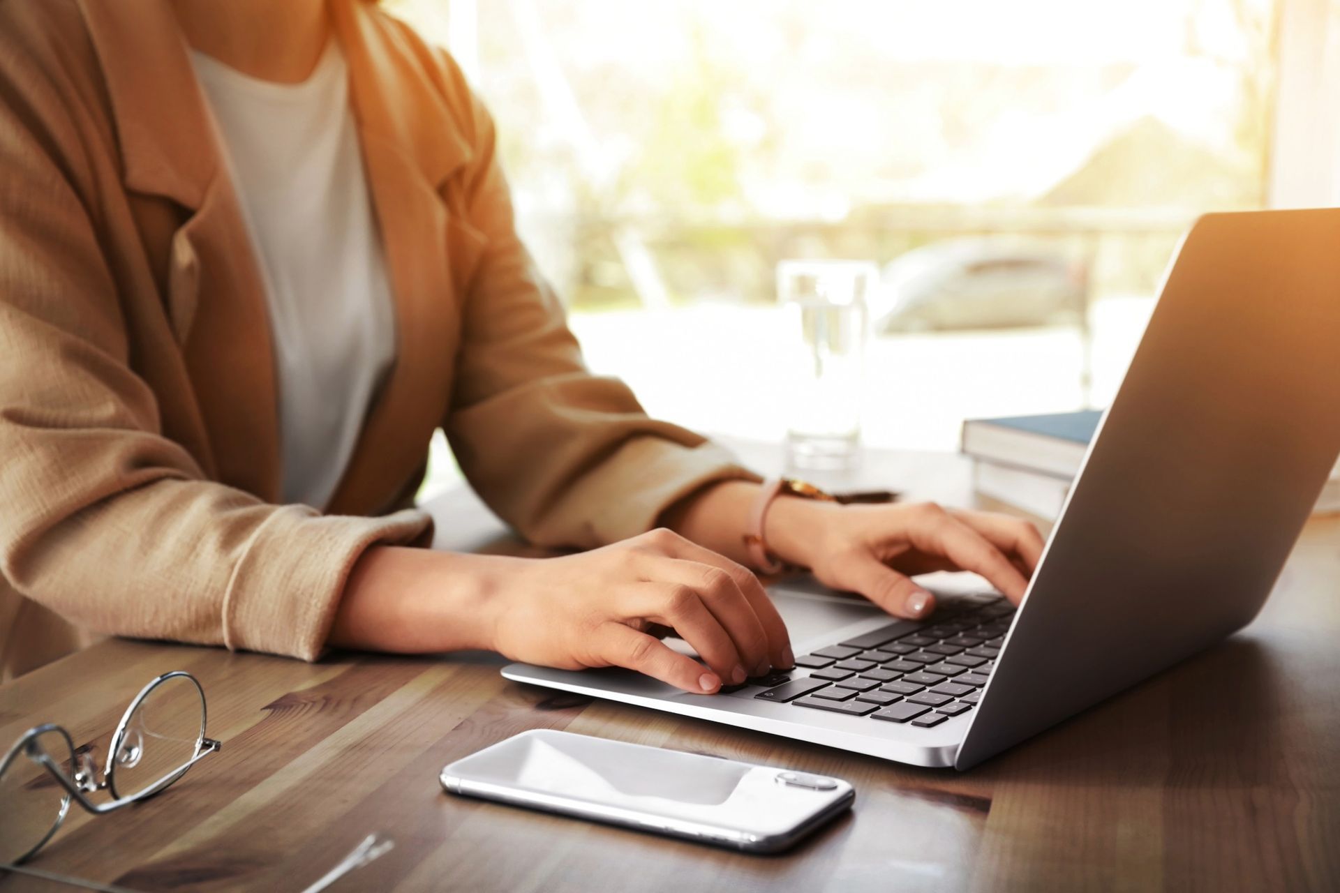 Woman typing on laptop at wooden desk near window, smartphone and glasses visible.