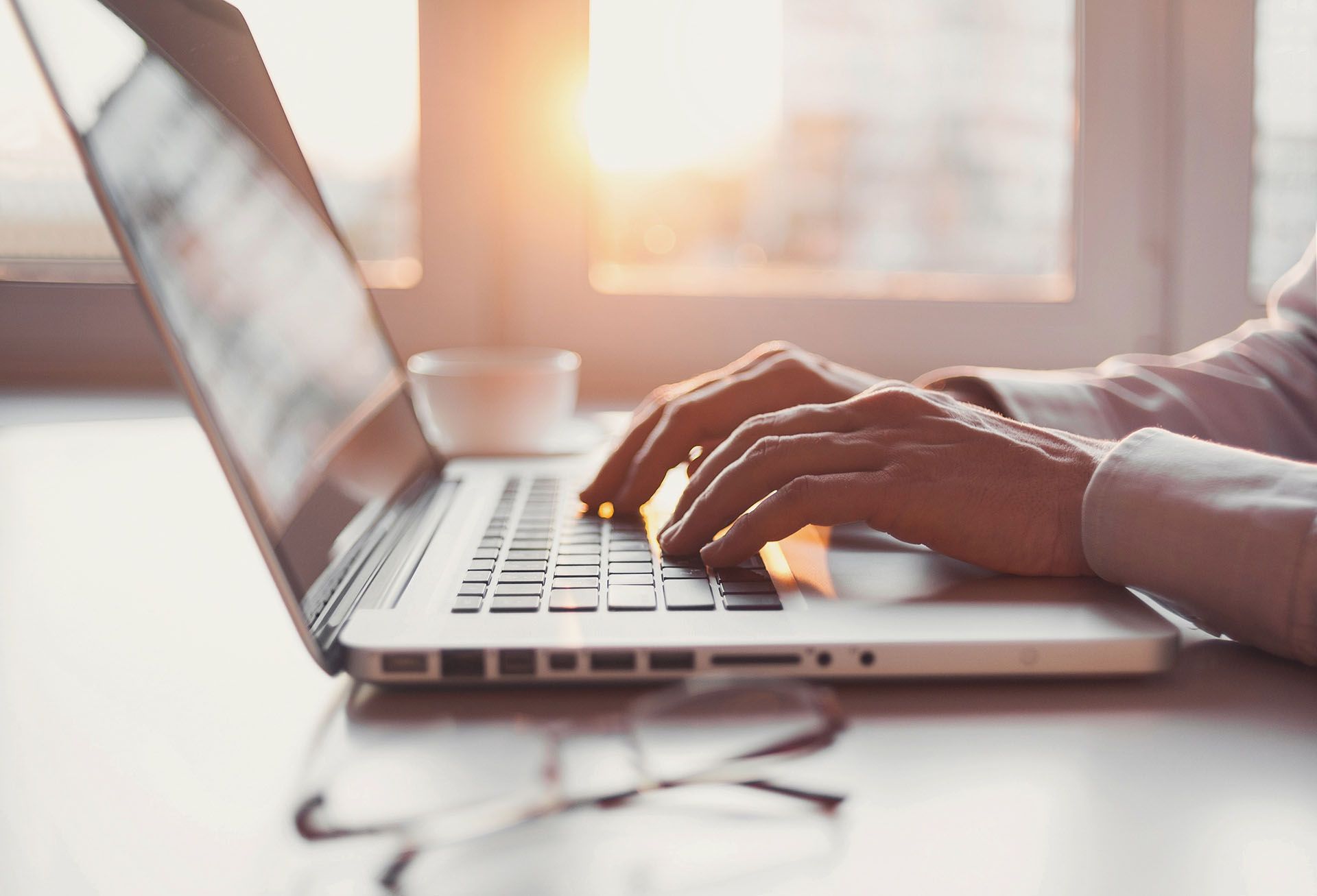 Hands typing on a laptop keyboard near a window with a bright sunset.