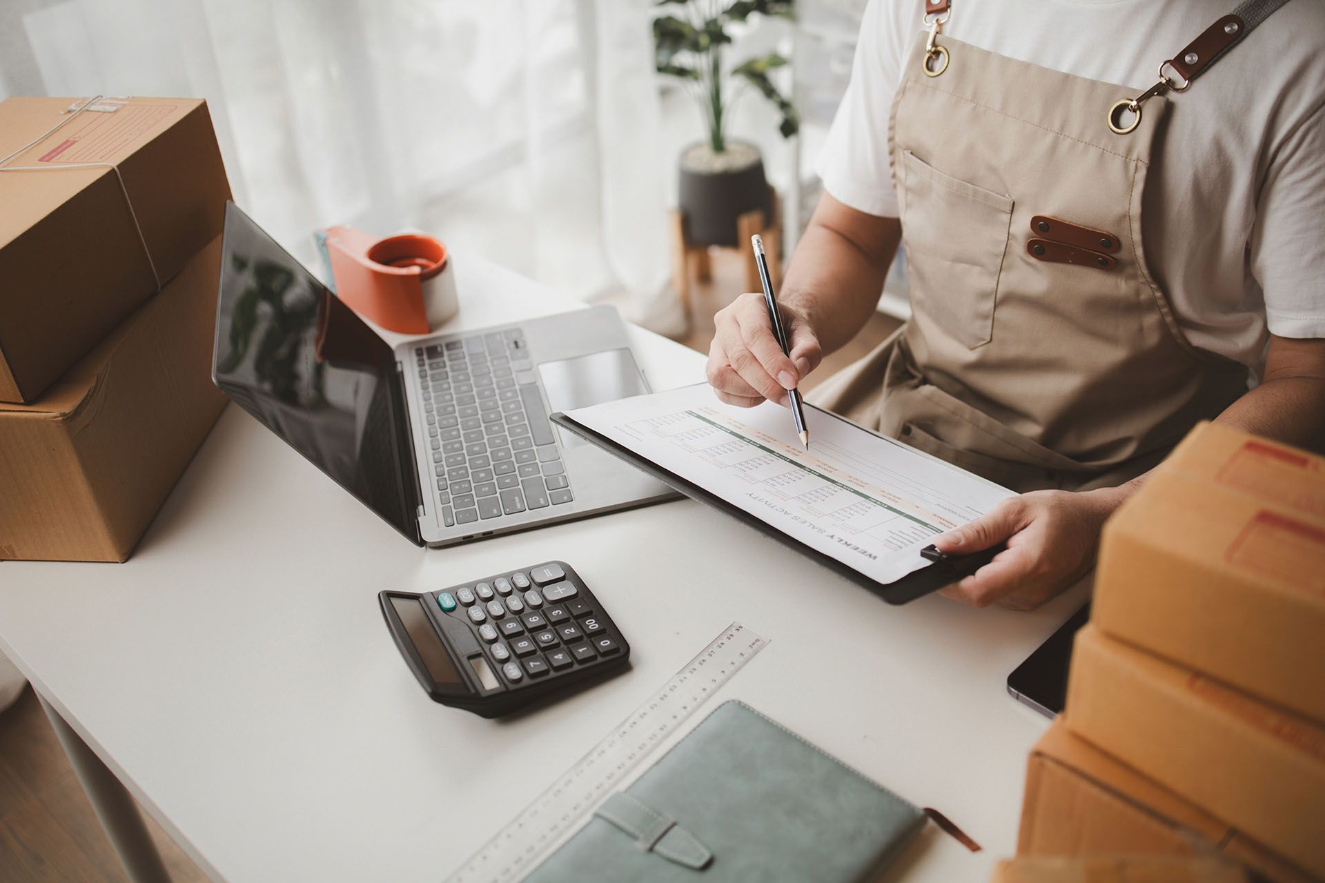 Person in apron reviews shipping paperwork at a desk, laptop open, surrounded by boxes.