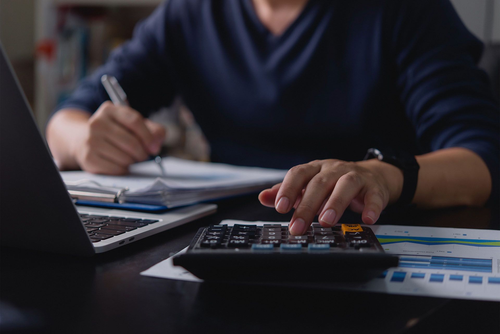 Person using calculator, writing notes, and working with laptop and charts at a desk.