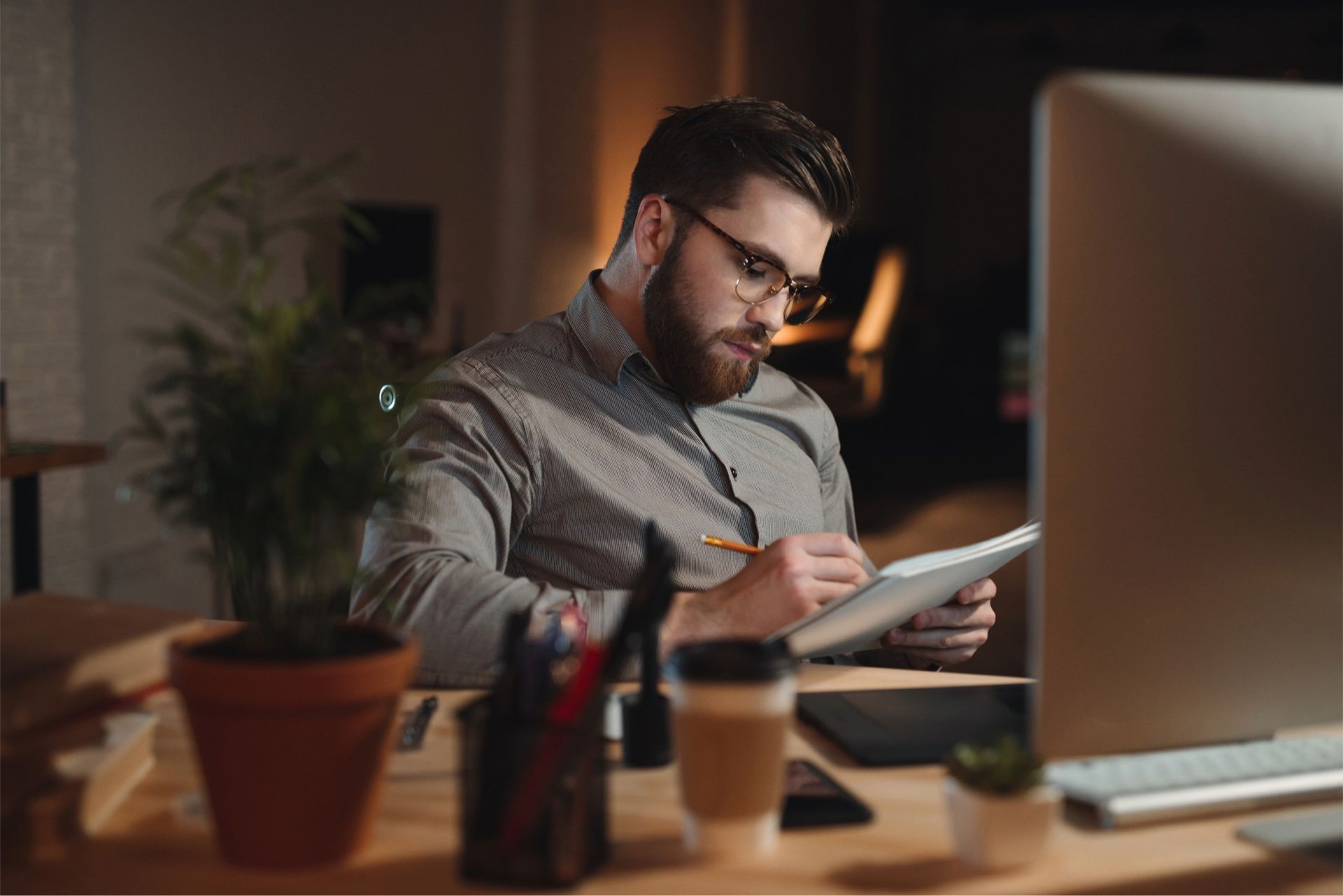 Man with glasses writing in notebook at desk with computer and coffee.
