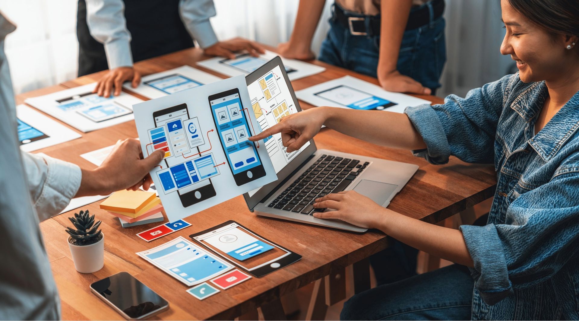 People collaborating on mobile app design, pointing at a laptop and printed wireframes on a wooden table.