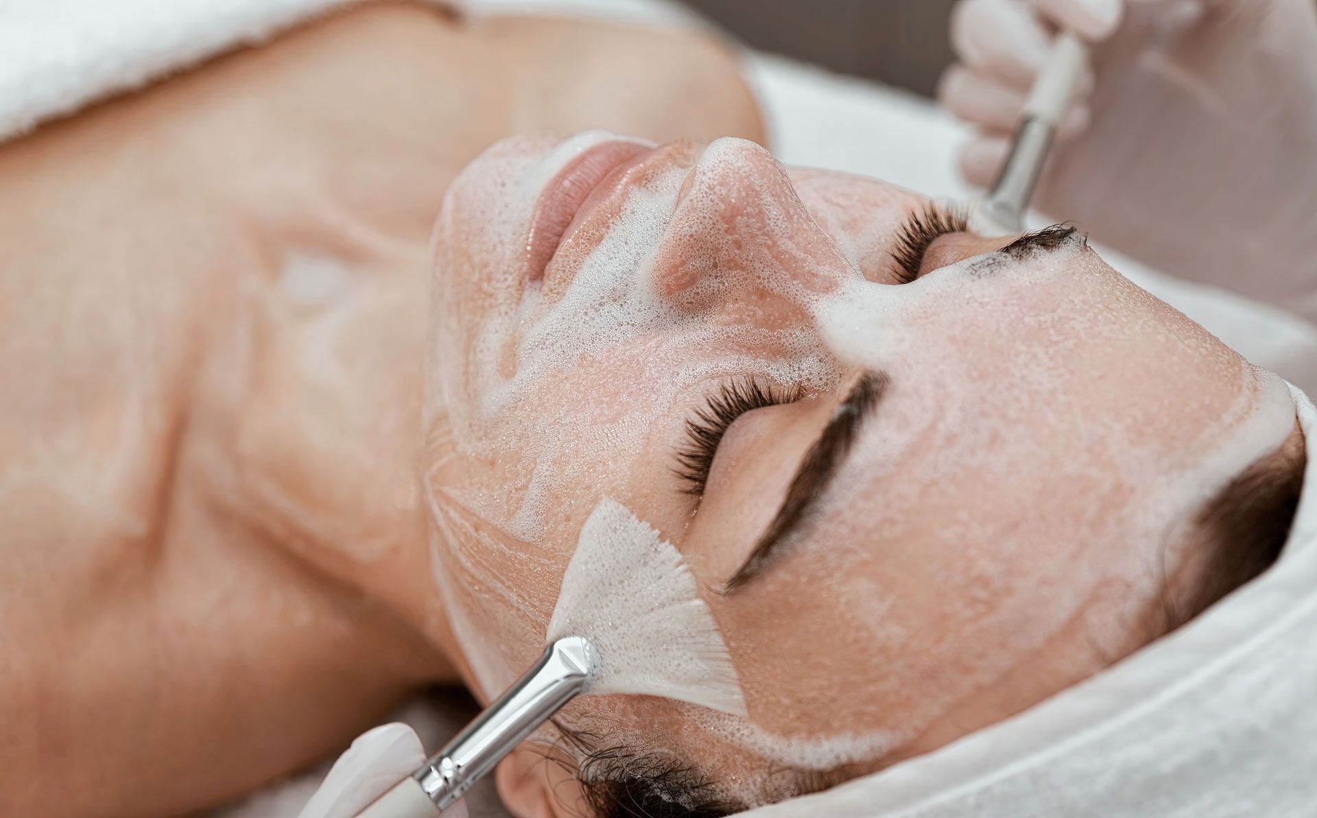 Person receiving a facial treatment; lying down with cream applied, tools held by gloved hands.