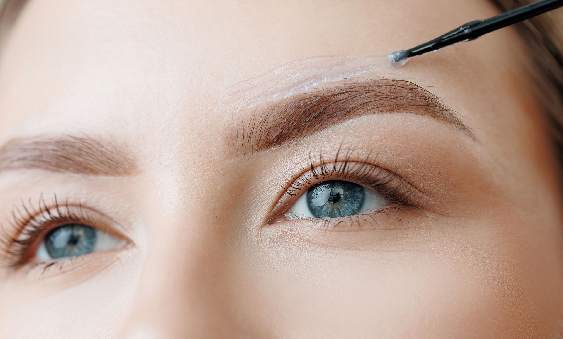 Woman applying eyebrow makeup with a brush. Blue eyes, close-up shot, cream-colored makeup.